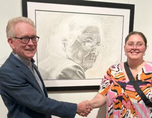A man in a blue suit shakes the hand of a young woman's in front of her artwork.